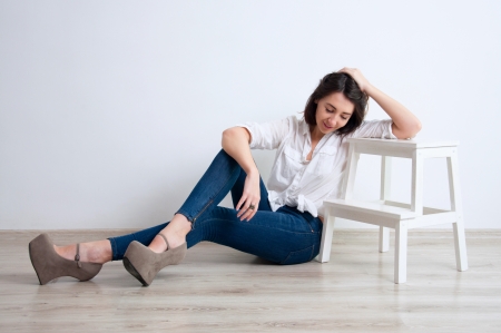 Pretty brunette woman wearing blue jeans, white shirt, stylish shoes, sitting on the floor in a minimalistic room against white wall, looking down, leaning against white stairs, smiling. Copy spaceの写真素材