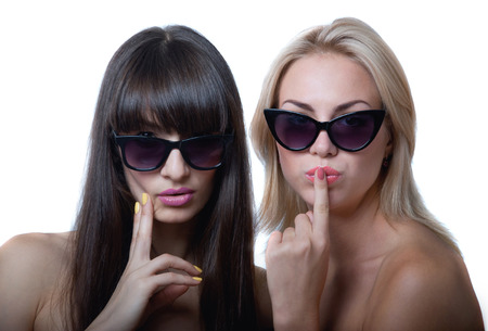 Studio portrait of two cute beautiful young women models wearing sun glasses, holding hands near lips and chin, smiling and looking at camera. Isolated on white backgroundの写真素材