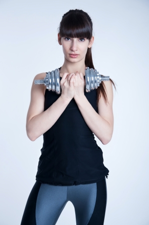Pretty young slim and fit brunette woman trainer holding a  heavy metal dumbbell, doing workout, training her muscles to become strong. Lookingat camera. In studio over gray backgroundの写真素材