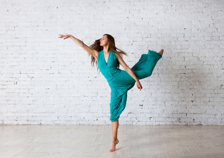Young beautiful woman dancer doing pirouette, posing, dancing, jumping in a studio, standing on a tiptoe, her blue turquoise long dress getting into drapes, hair flyong in the air. Over brick wall backgroundの写真素材