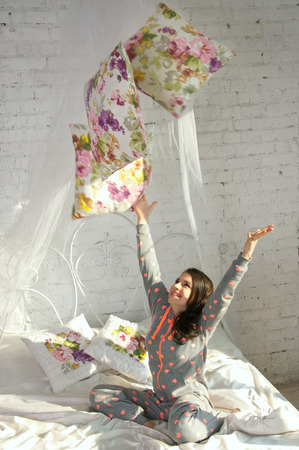 Pretty brunette teenage girl wearing pijamas, playing with pillows on a bed in the morning. Over brick wall backgroundの写真素材