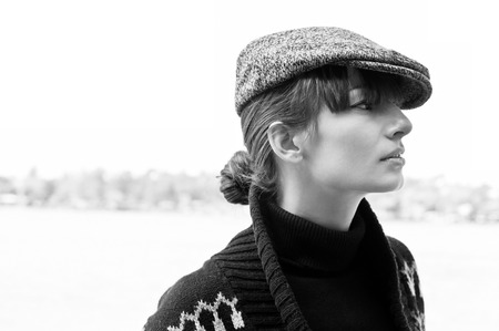 Black and white profile portrait of pretty young woman wearing black turtle-neck, boyish peaked cap and sweater, looking aside. High key portrait, copy spaceの写真素材