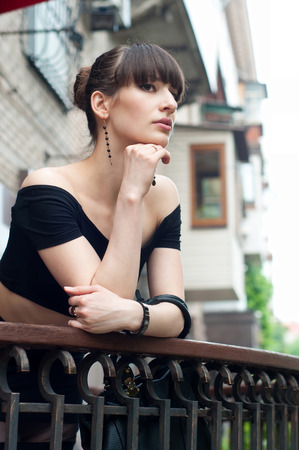 Pretty slim young brunette woman model wearing black off-the-shoulder top, elegant earrings, beautiful hairstyle, leaning over the balustrade of a balcony, looking somewhere, dreaming and enjoying the day.