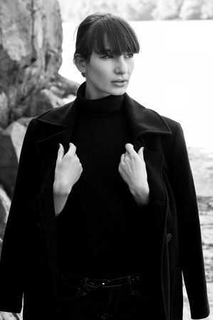 Elegant beautiful brunette woman model wearing tutpleneck sweater, holding her coat with hands, posing, looking aside, near rocks on a bright sunny day. Autumn fashion. High key portraitの写真素材