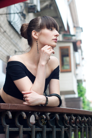 Pretty slim young brunette woman model wearing black off-the-shoulder top, elegant earrings, beautiful hairstyle, leaning over balustrade of a balcony, looking somewhere, dreaming and enjoying the dayの写真素材