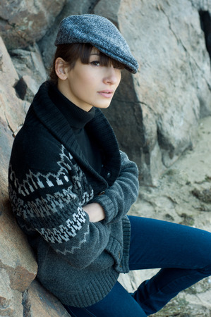Portrait of elegant beautiful brunette woman model wearing boyish clothes like turtleneck sweater, jacket, jeans, cap, red nails, siting near rocks, looking at camera. Autumn fashionの写真素材