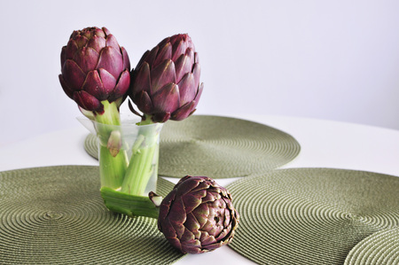 A bunch of beautiful fresh purple artichokes in a transparent jar on a table with round green table rugs, white background, copy spaceの写真素材