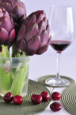 A bunch of beautiful fresh purple artichokes in a transparent jar on a table with round green table rugs, a glass of red wine at background, handful of ripe cherries in the front, white background, copy spaceの写真素材