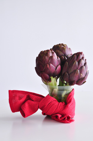 A bunch of beautiful fresh purple artichokes in a transparent jar on a white table, red kitchen towel forms a bow round a jar, white background, copy spaceの写真素材