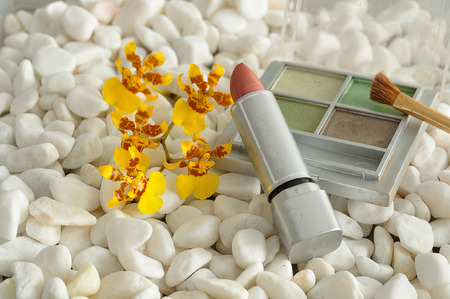 A palette with green shades of eye shadow and a brush, with lip stick displayed on white pebbles with a yellow orchidsの写真素材
