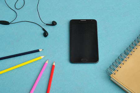 A slim phone with earphones displayed with a note book and coloring pencils on a blue backgroundの写真素材