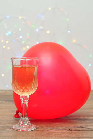 A glass of champagne and heart shape balloon against an out of focus light backgroundの写真素材
