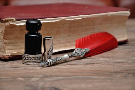 An old pen with a bottle of ink on a wooden table with a red bookの写真素材