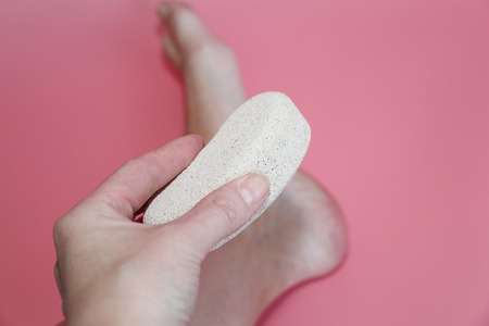 Pumice stone for foot care. In the photo a woman hand holds a pumice near the heel of her foot. Removal of rough skin on the heels of the feet with the help of pumice. Pink background.の写真素材