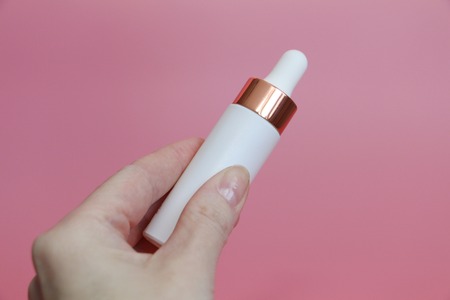 In the hand of a woman white tube with liquid. Female hand holds white bottle with elixir. Pink background.の写真素材