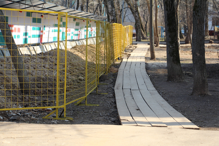 Construction is behind the fence. Around the construction paved the wooden road. Wooden road near the construction site. Nearby grow tall trees.の写真素材