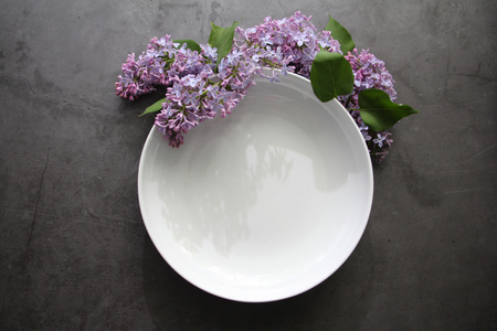 A bouquet of lilacs lies around the white plate. The white plate is empty. Background gray vintage. Vintage table. Lilac bouquet. Template for postcards.の写真素材