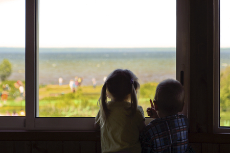 Two small children boy and girl look at the water and people from the window of the room in the summer.の写真素材