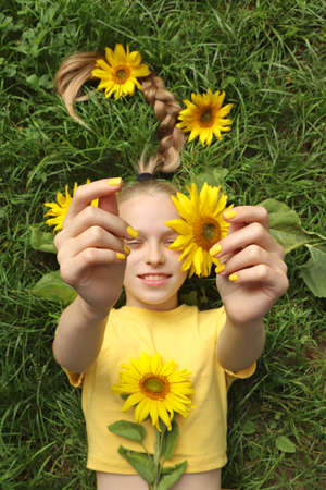 A beautiful girl with a yellow manicure lies on the grass with sunflowers.の写真素材
