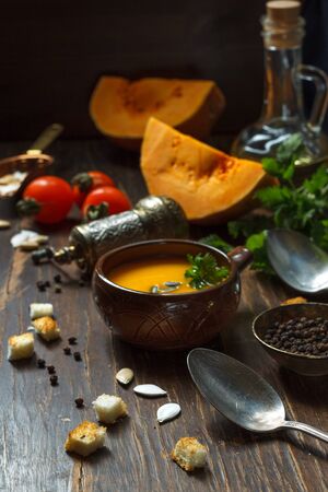 Homemade pumpkin soup in a brown pot , on a wooden kitchen table with pumpkin and tomatoes and olive oil, vertical orientationの写真素材