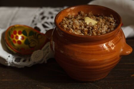 Buckwheat porridge in a brown ceramic pot with a piece of butter and a wooden painted spoon on a linen napkin, blurred backgroundの写真素材