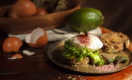 Healthy sandwich with poached egg, avocado and green salad on grain baguette on a copper plate, on a round wooden table with a dark backgroundの写真素材