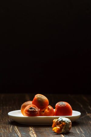 Rotten fruit with mold on a white saucer on a dark wooden table top with cracks, vertical with spaceの写真素材