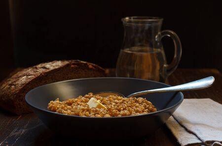 Useful porridge from a variety of wheat spelt in a gray plate with a piece of butter, rye bread and a jug of water in the backgroundの写真素材