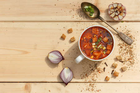 A Cup of rich soup with crackers and herbs. On a wooden table with a vintage spoon, a garlic head and a red onion. Horizontal orientation with space, top viewの写真素材