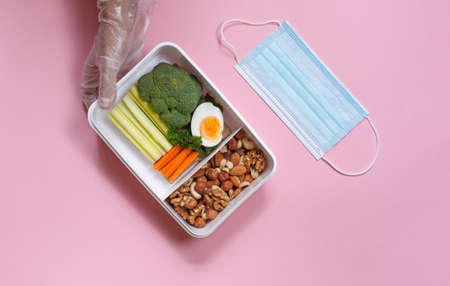 Hands in protective gloves with a useful lunch in a box and a protective mask. Horizontal top view...