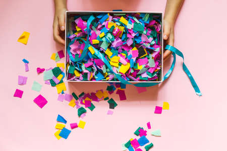Gift box with colored confetti in childrens hands. The view from the top on a pink surface,...
