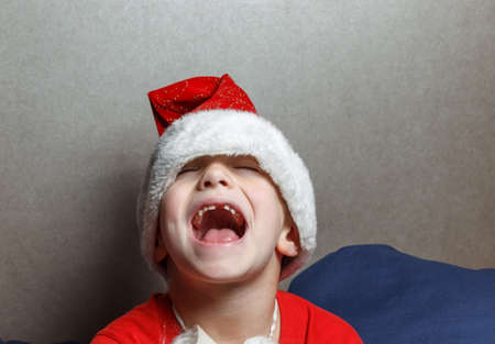 A little boy in a Santa hat with his eyes squeezed shut, laughing with his mouth wide open. Portrait on a brown backgroundの写真素材