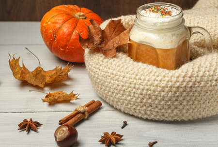 Pumpkin latte with thick milk foam and spices, in a glass mug, wrapped in a knitted white scarf. On a light wooden table with maple leaves and a pumpkinの写真素材