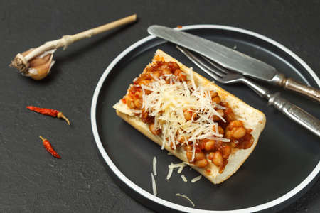 baked beans in sauce on a crispy toasted baguette with grated cheese. On a black background, on a...