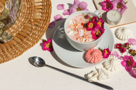 Cup with pink moon milk, with rosehip flowers, cinnamon, turmeric and other bright spices. On a white table with colored meringues, flowers, long spoon and a straw hatの写真素材