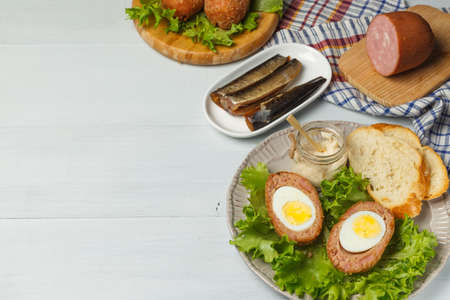 Scotch eggs on a plate with lettuce, toasted bread, and gravy. On a light gray wooden background with ham and sardines. Close-up, copy spaceの写真素材