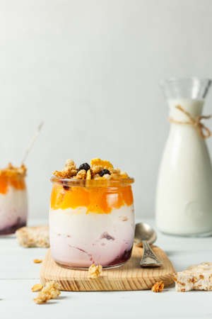 Homemade yogurt with fruit puree and granola, in a glass on a wooden stand, close-up. A jug of milk in the background.の写真素材