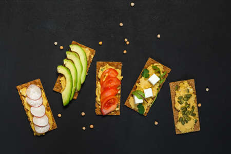 Rye crackers with hummus, avocado, tomatoes, radishes and tofu. Lean, vegetarian appetizer, vegetable sandwiches. On a black background, top view with spaceの写真素材