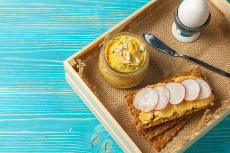 A wooden tray with a light, healthy breakfast. Boiled egg and rye crackers with hummus. Overhead, on a blue wooden background, with spaceの写真素材