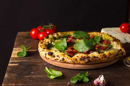Healthy Homemade vegetable pizza with spinach, pesto sauce and sun-dried tomatoes on a wooden round tray. black background, low key, horizontal, copy spaceの写真素材