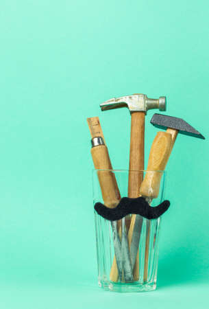 Vintage craft tools with wooden handles in a glass with a mock mustache pasted on. The concept of congratulations on father's Day, a men's holiday. on a green background, vertical with spaceの写真素材