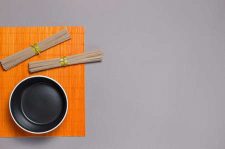 Two bundles of dried buckwheat soba noodles, on an orange bamboo napkin, and a black soup bowl....