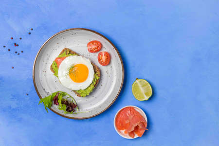 Healthy nutritious breakfast, toast on rye bread with avocado, salmon and egg. the sandwich is rich in protein and healthy fats. Top view on a blue backgroundの写真素材