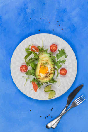 Original fried eggs baked in avocado, served with salad leaves, tomatoes and parmesan. A healthy, nutritious dish with a high protein content, superfood..Vertical frame, top, on a blue background with a fork and a knife , a copy of the spaceの写真素材