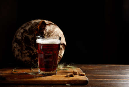 A glass with dark rye kvass or beer, with a thick foam. On a wooden board with crackers and spikelets. Rye round bread in the background. Horizontal, with space.の写真素材