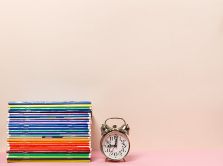 A stack of colorful notebooks and an alarm clock on the table. A horizontal frame with space. Pastel colors.の写真素材