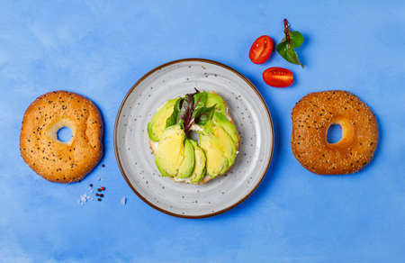 Bagel with avocado slices and salad leaves on a gray plate. On a blue background, horizontalの写真素材