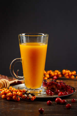 Hot or cold sea buckthorn juice, a useful vitamin drink to strengthen the immune system. A tall glass with sea buckthorn tea on a tray with a berry pie. Vertical frame on a black background. Copy of the spaceの写真素材