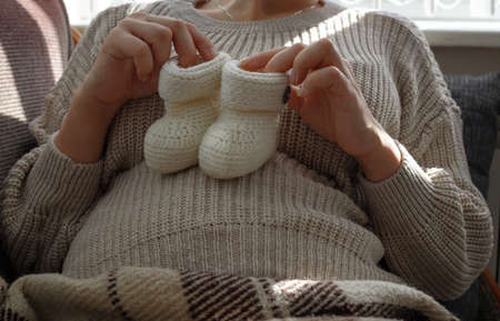Pregnant women in a beige sweater holds knitted baby white booties on her tummy