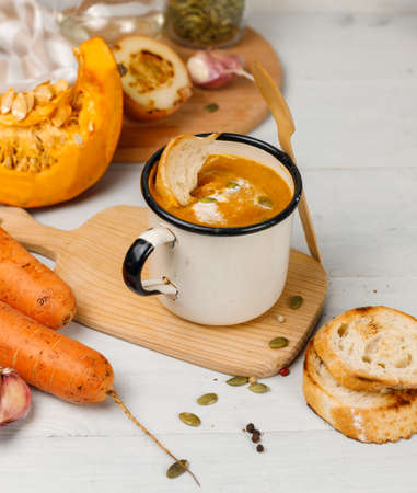Pumpkin soup with croutons in a metal mug, close-up on a light wooden table, with ingredients for cookingの写真素材